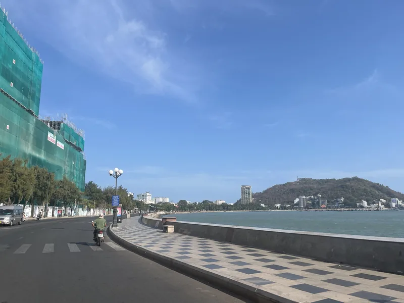 A checkered promenade lines the coastal road along Tran Phu Street in Vung Tau, with a hill and sea in the distance.